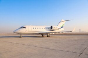 A white Bombardier Challenger 605 business jet parked on the ramp with a control tower visible in the background