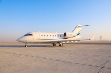 A white Bombardier Challenger 605 business jet parked on the ramp with a control tower visible in the background