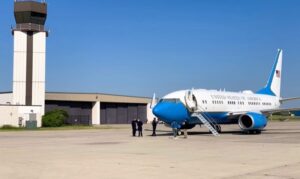 A blue-and-white aircraft marked United States of America parked on the ramp near an airport control tower with ground personnel nearby