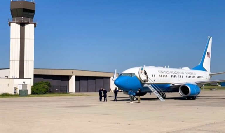 A blue-and-white aircraft marked United States of America parked on the ramp near an airport control tower with ground personnel nearby