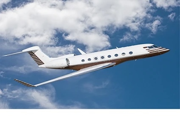 A white large-cabin business jet with a tan cheatline cruises against a blue, partly cloudy sky
